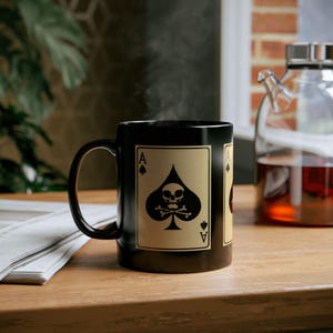 May include: Black ceramic mug featuring the Ace of Spades card design with a skull and crossbones. The card is tan with black details. Steam rises from the mug, suggesting a hot beverage. The mug sits on a wooden surface.