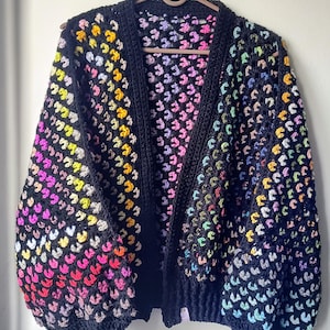 Può includere: Un cardigan in crochet nero con un motivo arcobaleno. Il cardigan ha un collo a V e maniche lunghe.