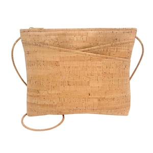 Puede incluir: Bolso bandolera de corcho marrón claro. Tiene forma rectangular, con cierre de cremallera y un bolsillo con solapa en la parte delantera. Cuenta con una fina correa ajustable. El bolso está sobre un fondo blanco.