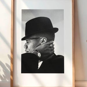 Puede incluir: Retrato enmarcado en blanco y negro de una persona con sombrero negro y gafas. El retrato se exhibe en una habitación con un mueble de madera, una lámpara y una planta en una cesta.