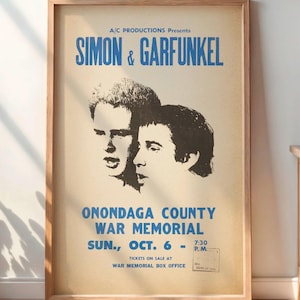 May include: A framed vintage poster for Simon & Garfunkel's concert at the Onondaga County War Memorial. The poster features the duo's portraits in black and white, with blue text indicating the venue and date. The poster is displayed in a room with a wooden cabinet and a potted plant.