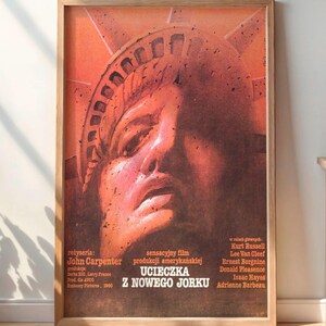 Puede incluir: Un póster de película enmarcado con un primer plano del rostro de la Estatua de la Libertad en tonos rojos y naranjas. El póster, en un marco de madera, se apoya en una pared blanca. El título está en polaco.