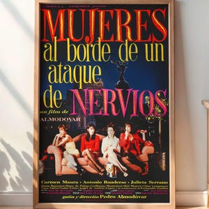 May include: A framed movie poster with the title "MUJERES al borde de un ataque de NERVIOS" in red and yellow lettering. The poster features a group of women and is displayed in a room with a wooden cabinet, a lamp, and a plant.