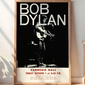 Op de afbeelding: Een ingelijste zwart-wit poster met Bob Dylan die gitaar speelt. De poster heeft de tekst "BOB DYLAN" in grote witte letters bovenaan en "CARNEGIE HALL FRIDAY OCTOBER 1 at 8:30 PM" onderaan. De poster zit in een houten lijst.