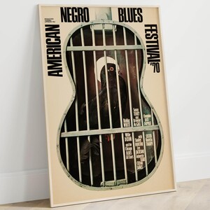 Puede incluir: Un póster enmarcado del Festival Americano de Blues Negro de 1970. El póster presenta una imagen en blanco y negro de una persona dentro de una jaula con forma de guitarra. El póster tiene texto en blanco y negro, incluyendo el nombre del festival y los nombres de los artistas.