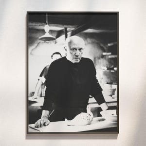 Könnte beinhalten: Ein schwarz-weiß gerahmtes Foto, das ein Porträt von Pablo Picasso in einem Atelier zeigt. Er trägt ein dunkles Langarmshirt und stützt seine Hände auf einen Tisch neben einem Keramikobjekt. Eine Hängelampe ist im Hintergrund zu sehen.