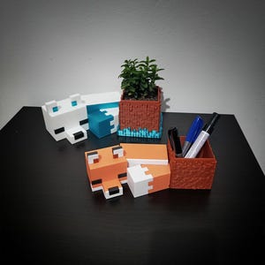 Könnte beinhalten: Eine Sammlung von Schreibtisch-Organizern und Pflanzgefäßen im Minecraft-Stil, 3D-gedruckt. Enthält einen weißen Eisbären, einen orangefarbenen Fuchs, einen braunen Pflanztopf mit einer kleinen grünen Pflanze und einen braunen Stifthalter.