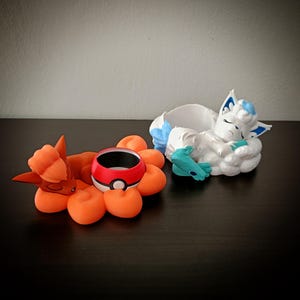 Op de afbeelding: Twee 3D-geprinte Pokémon-thema plantenbakken. Eén heeft een oranje vosachtig wezen met een rood-witte Poké Ball plantenbak. De andere is een wit vosachtig wezen met blauwe accenten, rustend in een wolkvormige plantenbak.