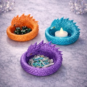 Puede incluir: Tres cuencos decorativos con forma de dragón en naranja, azul y morado. El cuenco naranja contiene dados, el cuenco azul contiene una vela y el cuenco morado contiene joyas. Cada cuenco tiene un diseño texturizado y escamado.