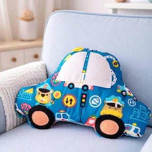 Può includere: Un cuscino blu a forma di auto con un finestrino bianco e ruote nere. Il cuscino presenta illustrazioni di agenti di polizia, semafori e paesaggi urbani. Il cuscino è su una poltrona azzurra.