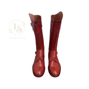 May include: A pair of red leather riding boots with a zip on the inside of each boot. The boots have a buckle strap at the top of the shaft and a buckle strap at the ankle.