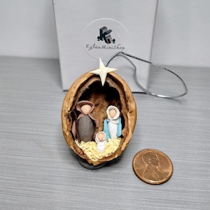 May include: A miniature nativity scene made from clay and placed inside a halved walnut shell. The figures are painted in brown, blue, and white. A small white star sits above the figures. The ornament is hanging from a silver string.