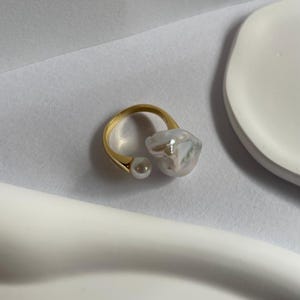 Peut inclure: Une bague en or avec deux perles de forme irrégulière. La bague est ouverte et les perles sont attachées à la bague de chaque côté.