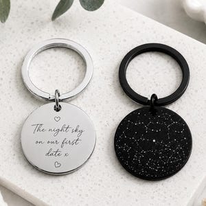 Könnte beinhalten: Zwei kreisförmige Schlüsselanhänger mit Metallringen. Einer ist silberfarben mit dem eingravierten Text 'The night sky on our first date x' und zwei kleinen Herzen. Der andere ist schwarz mit einer weißen Sternbildkarte auf der Oberfläche.