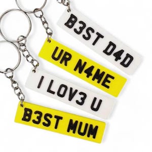May include: Four yellow and white keychains with black text. The keychains say "B3ST DAD", "UR N4ME", "I LOV3 U", and "B3ST MUM".