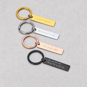 May include: Four rectangular metal keychains arranged vertically, each featuring a circular split ring. The keychains come in gold, silver, rose gold, and black finishes, with each rectangular tag engraved with a unique Morse code pattern.