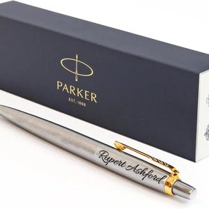 K&ouml;nnte beinhalten: Ein silberfarbener Kugelschreiber mit goldenem Clip und dem in schwarzer Schreibschrift eingravierten Namen Rupert Ashford. Der Stift liegt vor einer rechteckigen, dunkelblauen Parker-Geschenkbox mit Markenlogo und Text Est. 1888.