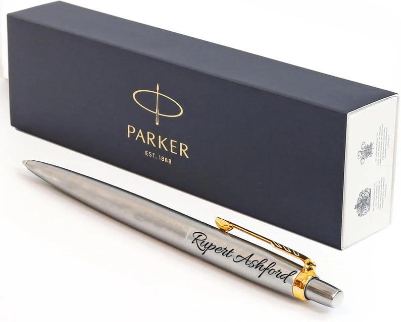 K&ouml;nnte beinhalten: Ein silberfarbener Kugelschreiber mit goldenem Clip und dem in schwarzer Schreibschrift eingravierten Namen Rupert Ashford. Der Stift liegt vor einer rechteckigen, dunkelblauen Parker-Geschenkbox mit Markenlogo und Text Est. 1888.
