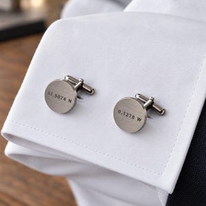 Puede incluir: Un par de gemelos redondos de color bronce cañón sujetos al puño de una camisa blanca. Un gemelo está grabado con el texto 51.5074 N y el otro con 0.1278 W, con un acabado metálico pulido y un cierre de palanca estándar en la parte posterior.