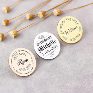 Könnte beinhalten: Drei runde Holz-Pins mit schwarzem Text und weißen Herzen. Die Pins zeigen "Best Man Ryan 12.03.2023", "Bridegroom Michelle 5.20.2023" und "Father of the Bride William 11.19.2023".