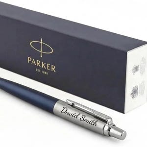 K&ouml;nnte beinhalten: Ein matter marineblauer Kugelschreiber mit einer silberfarbenen Kappe, auf der der Name David Smith eingraviert ist. Der Stift liegt vor einer rechteckigen, dunkelblauen Parker-Geschenkbox mit goldener Schrift und dem Text EST. 1888.