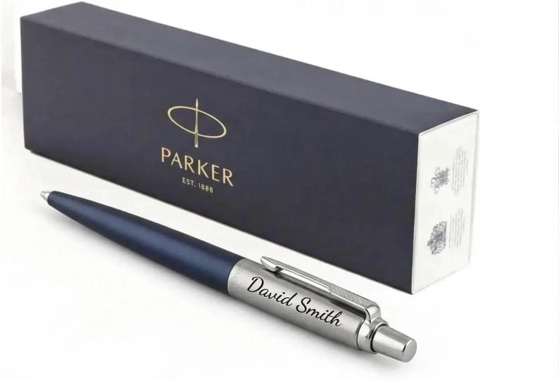 K&ouml;nnte beinhalten: Ein matter marineblauer Kugelschreiber mit einer silberfarbenen Kappe, auf der der Name David Smith eingraviert ist. Der Stift liegt vor einer rechteckigen, dunkelblauen Parker-Geschenkbox mit goldener Schrift und dem Text EST. 1888.