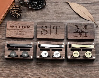 Op de afbeelding: Zes houten dozen met gegraveerde initialen en namen. Elke doos bevat een stropdasclip en manchetknopen. De dozen zijn gerangschikt in twee rijen van drie. De bovenste rij heeft de namen William, ST en Michael. De onderste rij heeft de initialen WD, ST en MT.