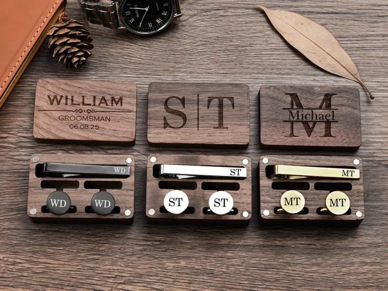 May include: Six wooden boxes with engraved initials and names. Each box contains a tie clip and cufflinks. The boxes are arranged in two rows of three. The top row has the names William, ST, and Michael. The bottom row has the initials WD, ST, and MT.
