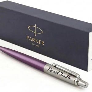 K&ouml;nnte beinhalten: Ein violetter Kugelschreiber mit einer silberfarbenen Kappe, auf der der Name David Smith eingraviert ist, vor einer dunkelblauen, rechteckigen Parker-Geschenkbox mit goldener Schrift und dem Aufdruck Parker Est. 1888 auf der Vorderseite.