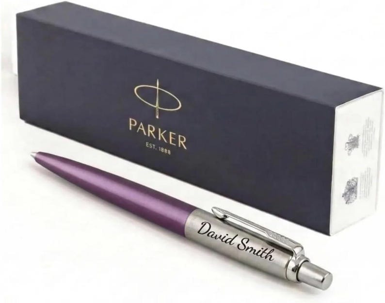 K&ouml;nnte beinhalten: Ein violetter Kugelschreiber mit einer silberfarbenen Kappe, auf der der Name David Smith eingraviert ist, vor einer dunkelblauen, rechteckigen Parker-Geschenkbox mit goldener Schrift und dem Aufdruck Parker Est. 1888 auf der Vorderseite.