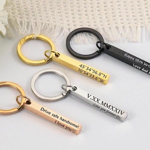 Könnte beinhalten: Drei goldene, silberne und schwarze Schlüsselanhänger mit gravierten Stäben. Der goldene Schlüsselanhänger hat den Text "Drive safe handsome! I love you" auf dem Stab graviert. Der silberne Schlüsselanhänger hat den Text "V.XX.MMXXIV Love you forever" auf dem Stab graviert. Der schwarze Schlüsselanhänger hat den Text "Happy 18th birthday Love dad xxx" auf dem Stab graviert. Der goldene Schlüsselanhänger hat auch Koordinaten auf dem Stab graviert: "45°34'56.8"N 10°54'33.6"W".