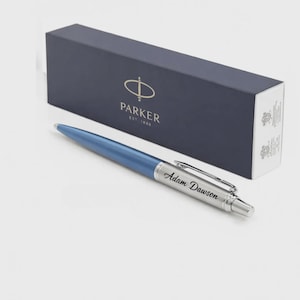K&ouml;nnte beinhalten: Ein blauer Kugelschreiber mit einem silberfarbenen Griffbereich, in den der Name Adam Dawson eingraviert ist, sowie einem silbernen Taschenclip. Der Stift liegt vor einer rechteckigen marineblauen Parker-Geschenkbox mit goldfarbenem Text.