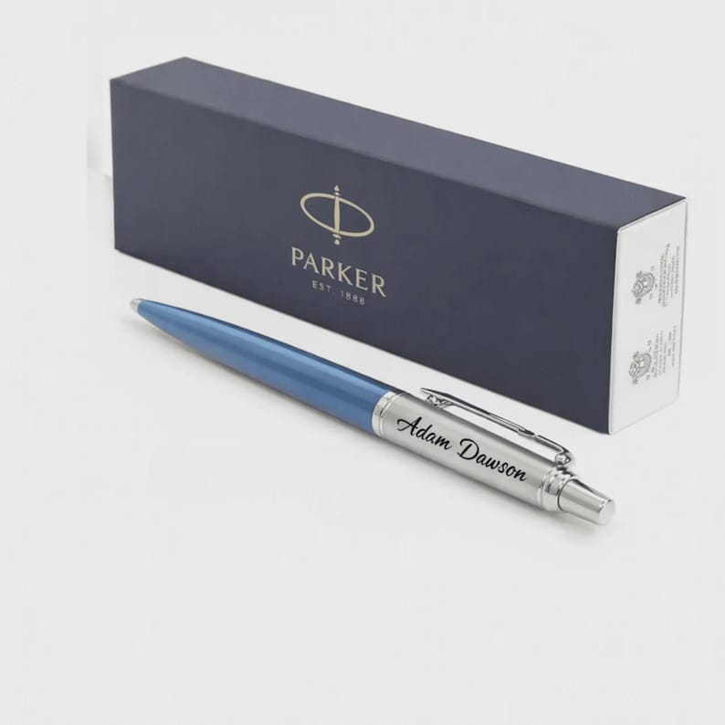 K&ouml;nnte beinhalten: Ein blauer Kugelschreiber mit einem silberfarbenen Griffbereich, in den der Name Adam Dawson eingraviert ist, sowie einem silbernen Taschenclip. Der Stift liegt vor einer rechteckigen marineblauen Parker-Geschenkbox mit goldfarbenem Text.