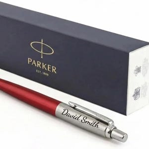 K&ouml;nnte beinhalten: Ein roter und silberner Kugelschreiber mit dem eingravierten Namen David Smith auf dem silbernen Schaft. Der Stift liegt vor einer rechteckigen, dunkelblauen Parker-Geschenkbox mit dem goldenen Parker-Logo und dem Text EST. 1888.