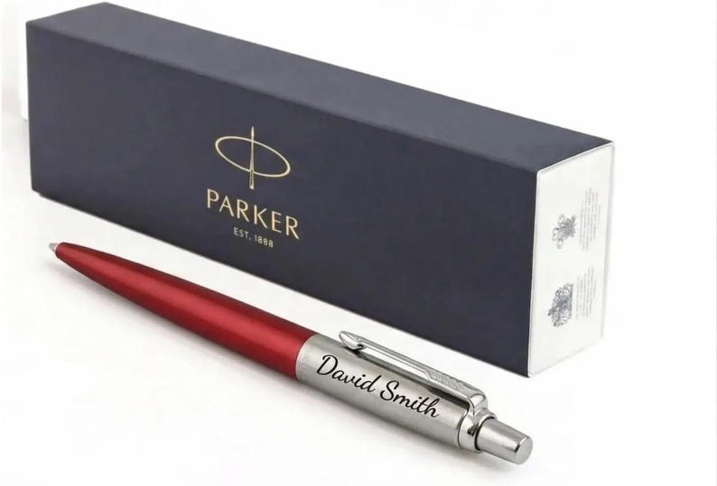 K&ouml;nnte beinhalten: Ein roter und silberner Kugelschreiber mit dem eingravierten Namen David Smith auf dem silbernen Schaft. Der Stift liegt vor einer rechteckigen, dunkelblauen Parker-Geschenkbox mit dem goldenen Parker-Logo und dem Text EST. 1888.