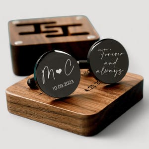 May include: A pair of black cufflinks with engraved designs. One cufflink says "M&C" with a heart and the date "10.09.2023". The other cufflink says "Forever and always". The cufflinks are displayed on a wooden box.