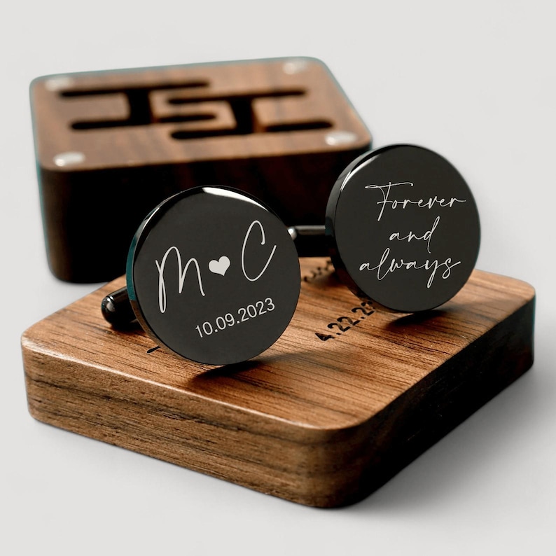 May include: A pair of black cufflinks with engraved designs. One cufflink says "M&C" with a heart and the date "10.09.2023". The other cufflink says "Forever and always". The cufflinks are displayed on a wooden box.