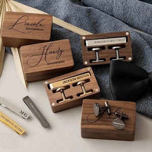 May include: Wooden boxes with engraved names and the word "Groom" on the lid. Each box contains cufflinks or a tie clip. The boxes are made of dark wood with a natural grain.