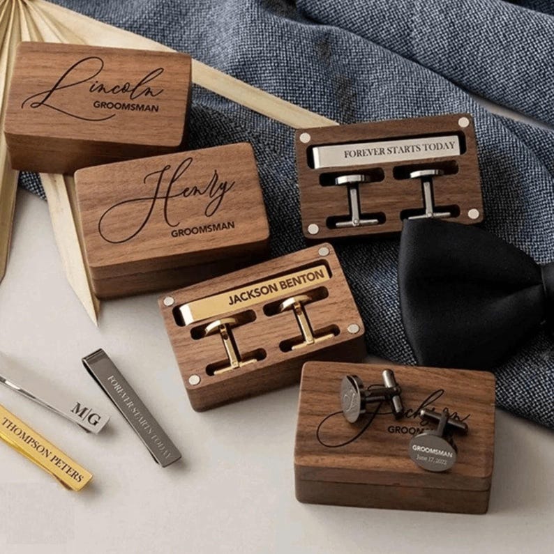 May include: Wooden boxes with engraved names and the word "Groom" on the lid. Each box contains cufflinks or a tie clip. The boxes are made of dark wood with a natural grain.