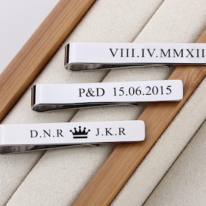 May include: Three silver tie clips with engraved text. The top clip reads "VIII.IV.MMXII", the middle clip reads "P&D 15.06.2015", and the bottom clip reads "D.N.R. J.K.R." with a crown symbol between the initials.