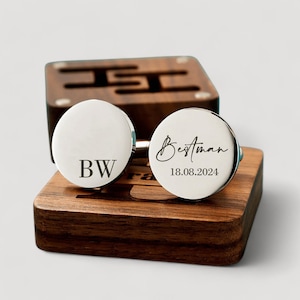 May include: A pair of silver cufflinks with engraved text. One cufflink says "Bestman" with the date "18.08.2024" and the other cufflink says "BW". The cufflinks are displayed on a wooden box.