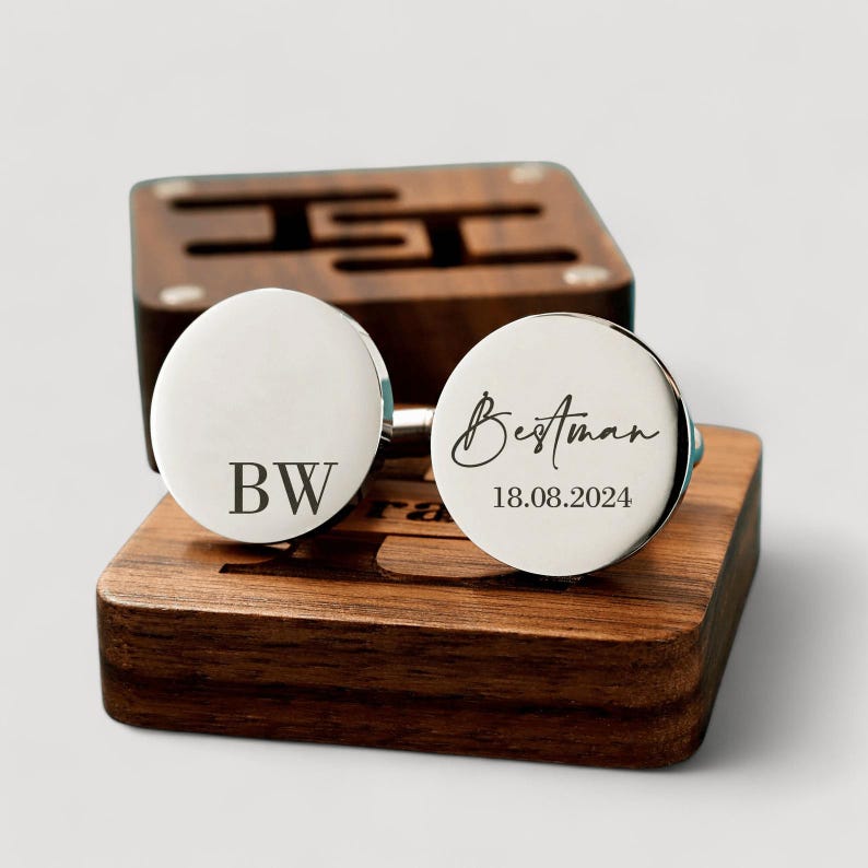 May include: A pair of silver cufflinks with engraved text. One cufflink says "Bestman" with the date "18.08.2024" and the other cufflink says "BW". The cufflinks are displayed on a wooden box.