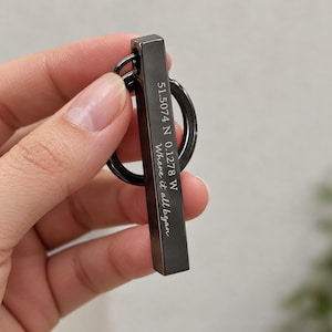 May include: A black metal bar keychain featuring a circular ring attachment. The bar is engraved with the coordinates 51.5074 N 0.1278 W and the phrase Where it all began in a white script font, creating a contrast against the dark surface.