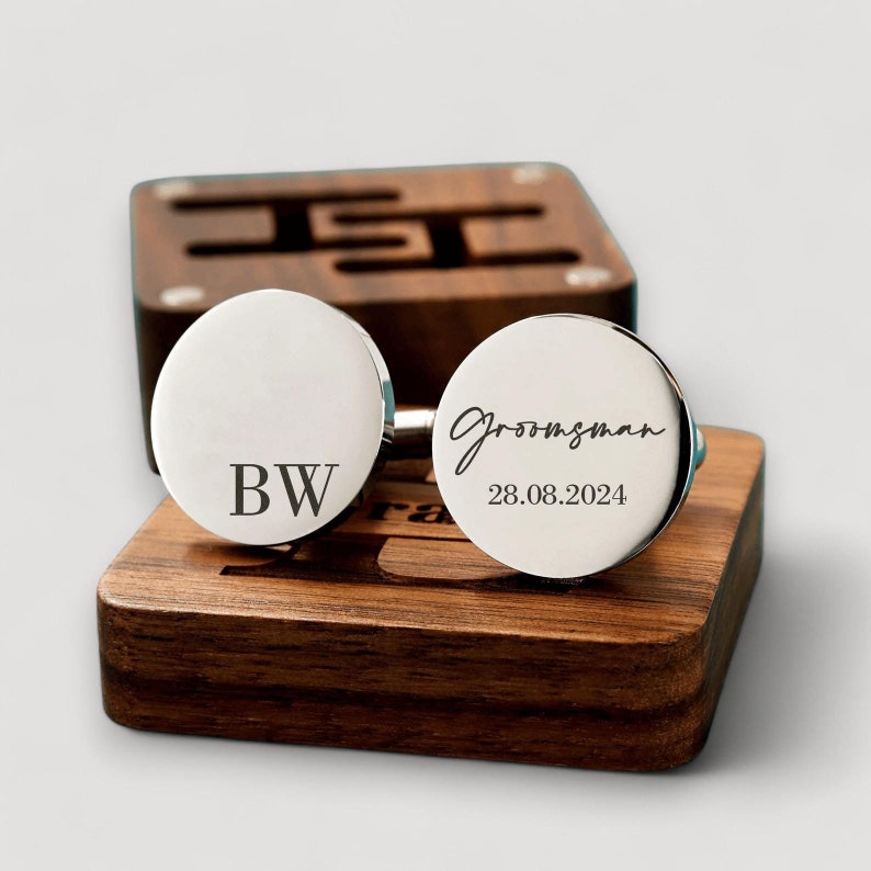 May include: A pair of silver cufflinks with engraved text. One cufflink says "BW" and the other says "Groomsman 28.08.2024". The cufflinks are displayed on a wooden box.