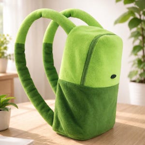 May include: A plush green backpack with a two-tone design. The top portion is a lighter green, while the bottom is a darker shade. It features a zipper closure and two padded shoulder straps. A small black oval is on the front.