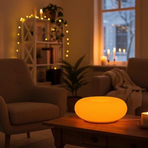 May include: An illuminated, orange-colored, oval-shaped lamp sits on a wooden coffee table. The lamp emits a warm glow, illuminating the surrounding living room. The room features a neutral color palette with a cozy armchair and a lit candle.