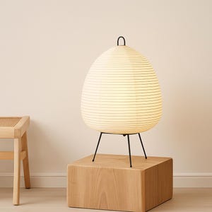 May include: A modern egg-shaped table lamp with a paper shade and black metal legs. The lamp is illuminated, casting a warm glow. It sits on a wooden cube, and the design is minimalist and contemporary.