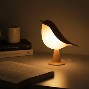 May include: A wooden bird-shaped table lamp with a glowing white body and a dark brown head and wings. The lamp sits on a wooden base and is plugged into a wall outlet. A white mug and books are in the background.