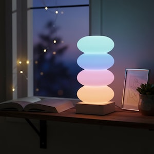 May include: A modern, stacked stone-shaped lamp with a soft, colorful glow. The lamp emits a gradient of colors, including light blue, purple, pink, and yellow. It sits on a white base, next to a framed picture and a potted plant.