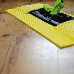 Puede incluir: Un mocho amarillo y negro con un mango verde se muestra en un suelo de madera. La cabeza del mocho está hecha de un material suave y absorbente.